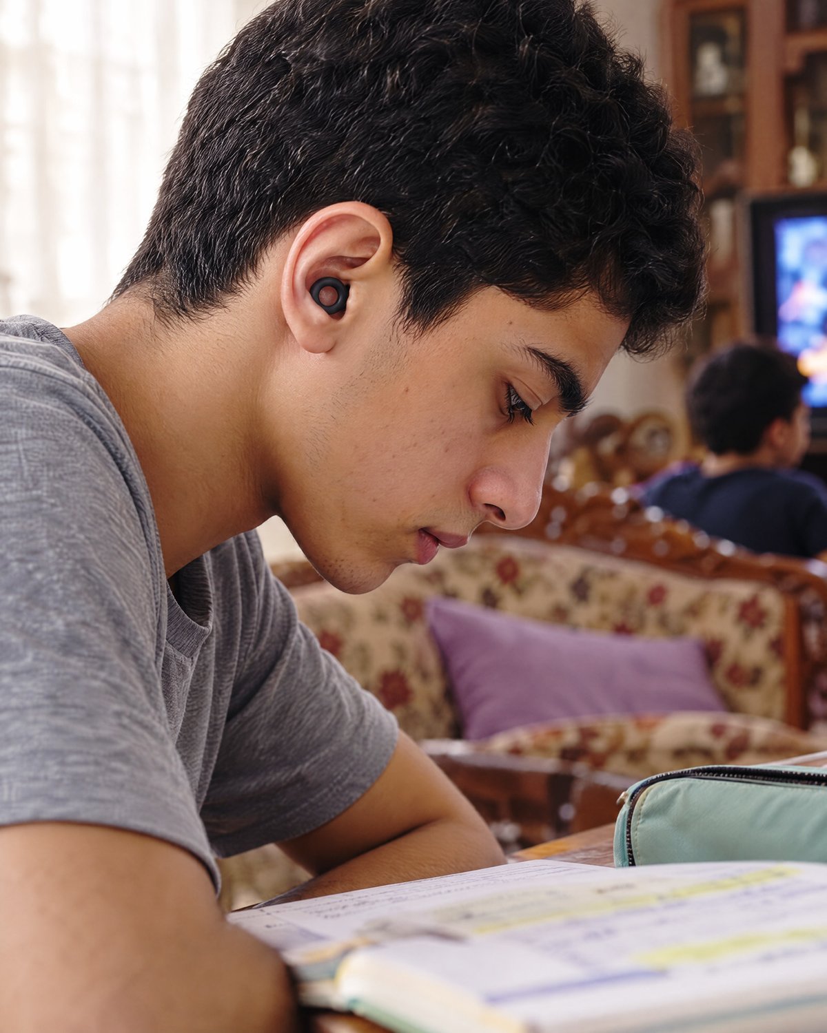 A teenage boy studying at home, wearing a Black PLUGS earplug while the TV plays behind him