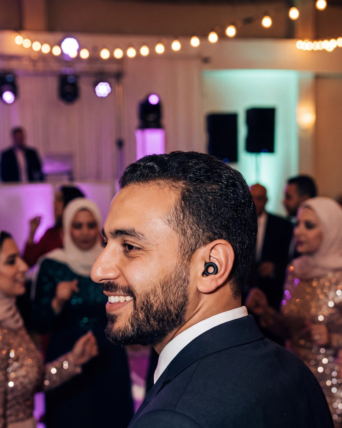 A smiling man at a wedding party, wearing a Black PLUGS earplug